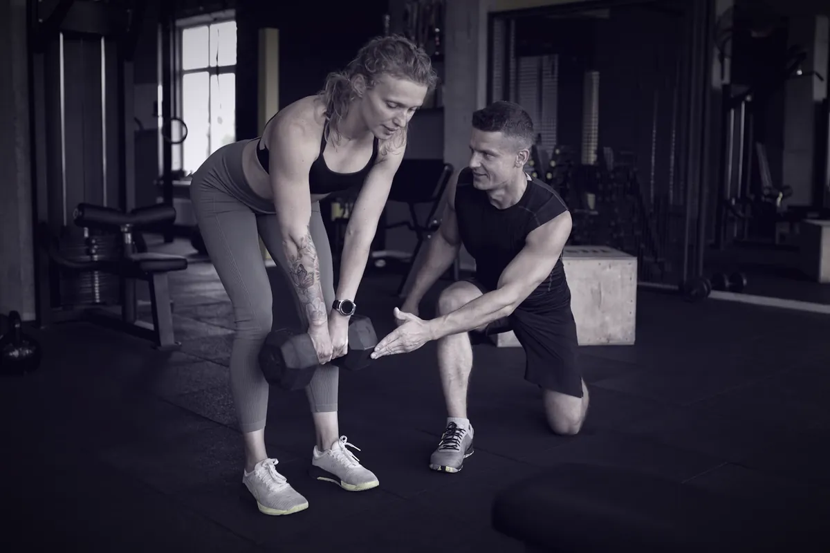 Personal trainer ajoelhado corrigindo a pegada de uma aluna que segura um halter, numa academia.